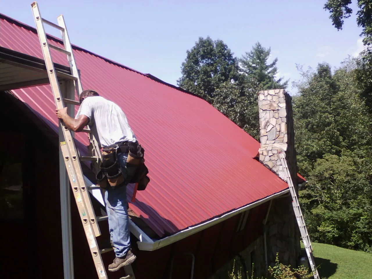 Roofer on a ladder beside a red metal roof and stone chimney — Soffit & Fascia Repair by Roofing Brothers in Trinity Center